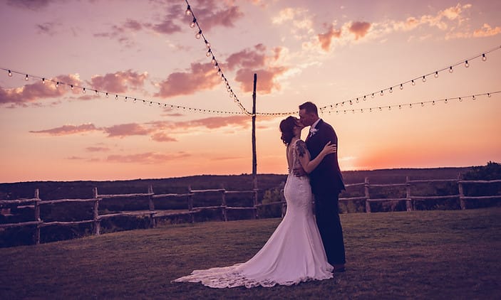 A bride and groom kiss and embrace in front of a sunset on their wedding day